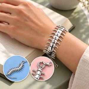 Pode incluir: Uma pulseira prateada em forma de centopeia, com partes do corpo segmentadas e múltiplas pernas pequenas. A pulseira possui um fecho lagosta e uma corrente extensora ajustável, exibida num pulso e em dois detalhes circulares.