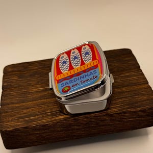 May include: A small, hinged metal tin with a colourful sardine design. The lid features three fish illustrations and the text "SARDINHAS em tomate." The tin is open, revealing its empty interior. It rests on a dark wooden surface.