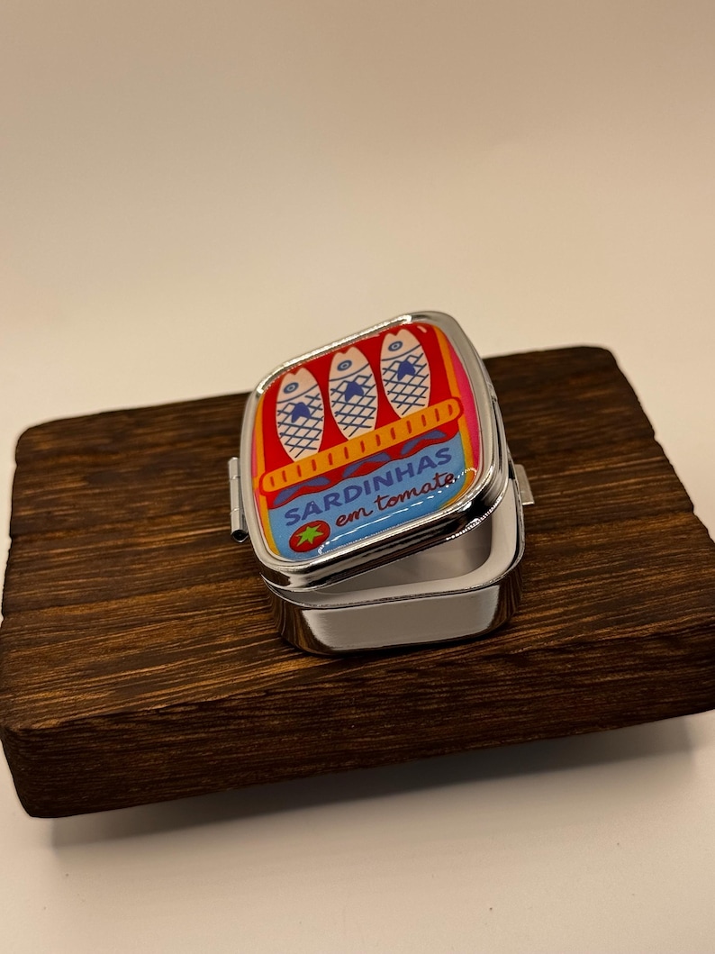 May include: A small, hinged metal tin with a colourful sardine design. The lid features three fish illustrations and the text "SARDINHAS em tomate." The tin is open, revealing its empty interior. It rests on a dark wooden surface.