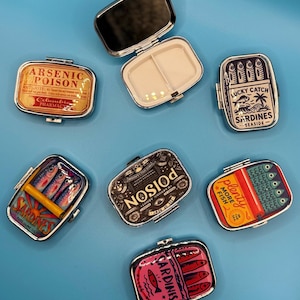 May include: Several small, rectangular metal pill cases with different designs. One is open, showing a mirror and a divided interior. The other cases have sardine and poison-themed graphics, with text including "ARSENIC POISON" and "SARDINES".