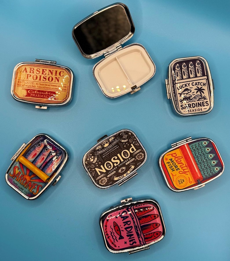 May include: Several small, rectangular metal pill cases with different designs. One is open, showing a mirror and a divided interior. The other cases have sardine and poison-themed graphics, with text including "ARSENIC POISON" and "SARDINES".