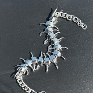 Pode incluir: Uma pulseira de metal prateado em forma de centopeia, apresentando um corpo segmentado com múltiplas pernas salientes ao longo do seu comprimento. A peça inclui uma secção de corrente de elos em ambas as extremidades e um fecho lagosta.