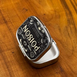 May include: A small, open, silver metal tin with a black lid. The lid displays the word "POISON" in white, along with floral and skull motifs. Text includes "Warning About". The tin sits on a wooden surface.
