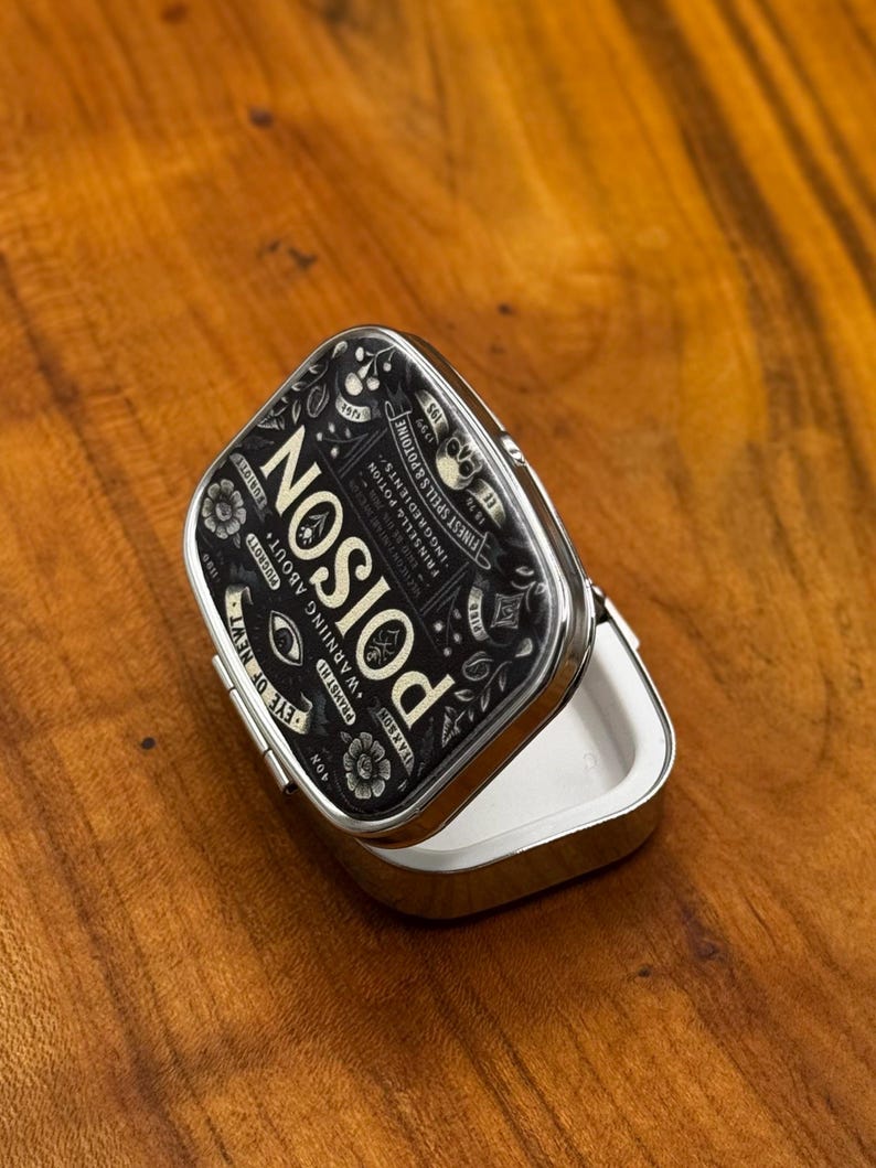 May include: A small, open, silver metal tin with a black lid. The lid displays the word "POISON" in white, along with floral and skull motifs. Text includes "Warning About". The tin sits on a wooden surface.