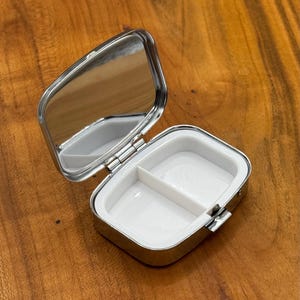 May include: A small, open, silver pill box with a mirror on the inside of the lid. The interior is white and divided into two compartments. The box is rectangular with rounded corners and a hinged lid.