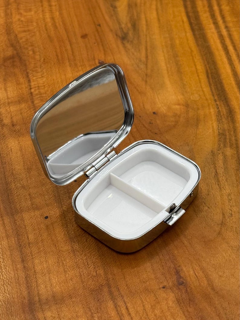 May include: A small, open, silver pill box with a mirror on the inside of the lid. The interior is white and divided into two compartments. The box is rectangular with rounded corners and a hinged lid.