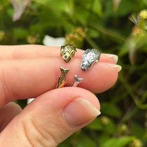 Può includere: Un anello aperto con quattro piccole figure di pesci in tonalità alternate oro e argento. I pesci sono disposti in un motivo circolare, con due pesci più grandi rivolti l'uno verso l'altro e due pesci più piccoli posizionati tra loro.