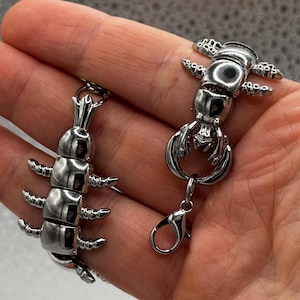 Może przedstawiać: Dwa srebrne wisiorki w kształcie owadów. Jeden to wzór stonogi, a drugi to wzór homara. Oba mają skomplikowane detale i są przymocowane do zapięcia.