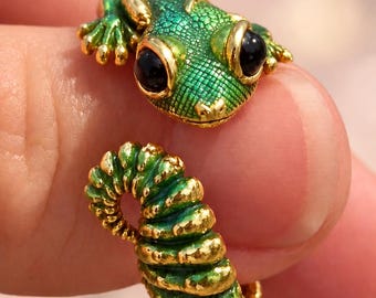 May include: A gold-toned open ring shaped like a lizard with a textured green enameled body and head. The lizard features two large, smooth black circular eyes and a segmented, curled tail that forms the band of the ring.