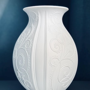 May include: A matte white ceramic vase featuring a rounded body and a flared rim. The surface is decorated with raised, embossed swirling patterns and vertical lines that create a textured relief across the exterior of the vessel.