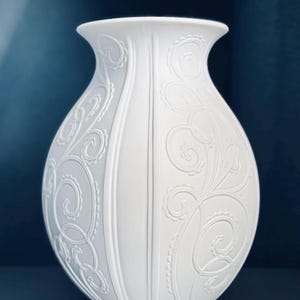 Könnte beinhalten: Eine matte, weiße Keramikvase mit bauchigem Körper und ausgestelltem Rand. Die Oberfläche ist mit erhabenen, geprägten Schnörkeln und Wirbelmustern verziert, die sich um das Äußere winden und durch vertikale, glatte Vertiefungen getrennt sind.