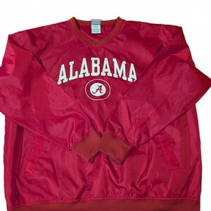May include: A red long-sleeve pullover windbreaker featuring a V-neck collar and ribbed cuffs. The word ALABAMA is printed in white capital letters across the chest, positioned above a white circular emblem containing a red letter A.