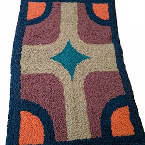 May include: A rectangular tufted rug featuring a geometric pattern with a central teal diamond shape surrounded by beige, purple, and dark blue sections. The four corners of the rug are accented with bright orange semi-circular shapes.