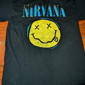 May include: A black short-sleeve t-shirt featuring the word NIRVANA in distressed blue lettering across the chest. Below the text is a large yellow smiley face graphic with black X eyes, a wavy mouth, and a tongue sticking out to the right side.
