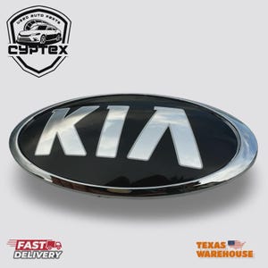 May include: Black and chrome Kia emblem. The oval emblem features the word "KIA" in silver, set against a black background. The emblem is surrounded by a chrome border. The image also includes the text "Used Auto Parts" and "Cyptex".