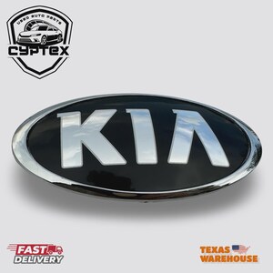 May include: A black and chrome Kia emblem with the word "KIA" in silver lettering. The emblem is oval-shaped and has a shiny chrome border. The image also includes the text "FAST DELIVERY" and "TEXAS WAREHOUSE".