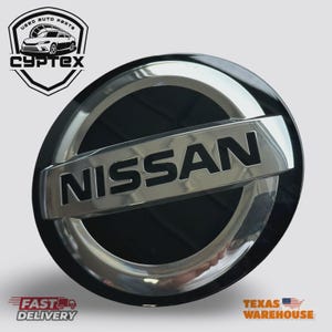 May include: A black and chrome Nissan emblem. The word "NISSAN" is prominently displayed in silver lettering across the center. The emblem is circular with a shiny chrome border. The top left corner features a logo with the text "CYPTEX" and "USED AUTO PARTS".