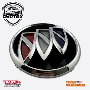 May include: A chrome-finished Buick emblem with a black background and three colored shield shapes. The shields are red, silver, and blue. The emblem is oval-shaped. The image also includes the text "CYPTEX" and "USED AUTO PARTS".