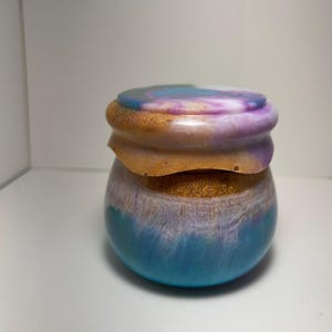 May include: A decorative jar with a lid. The jar is a gradient of colors, including blue, purple, gold, and white. The lid is a swirl of blue, purple, and white. The jar has a rounded shape.