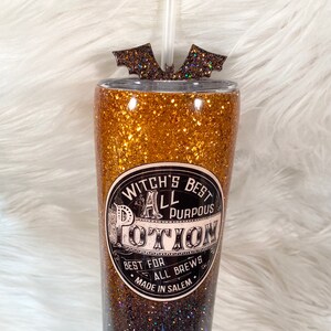 May include: A glitter-covered tumbler with a straw and bat-shaped topper. The tumbler features a gold and black ombre design and a label that reads "Witch's Best All Purpous Potion". The text "Best for All Brews, Made in Salem" is also on the label.