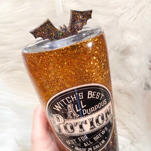 May include: A glittery tumbler with a bat-shaped topper and a straw. The tumbler is gold and black with the text "Witch's Best All Purpous Potion". The design is perfect for Halloween or a witchy theme.