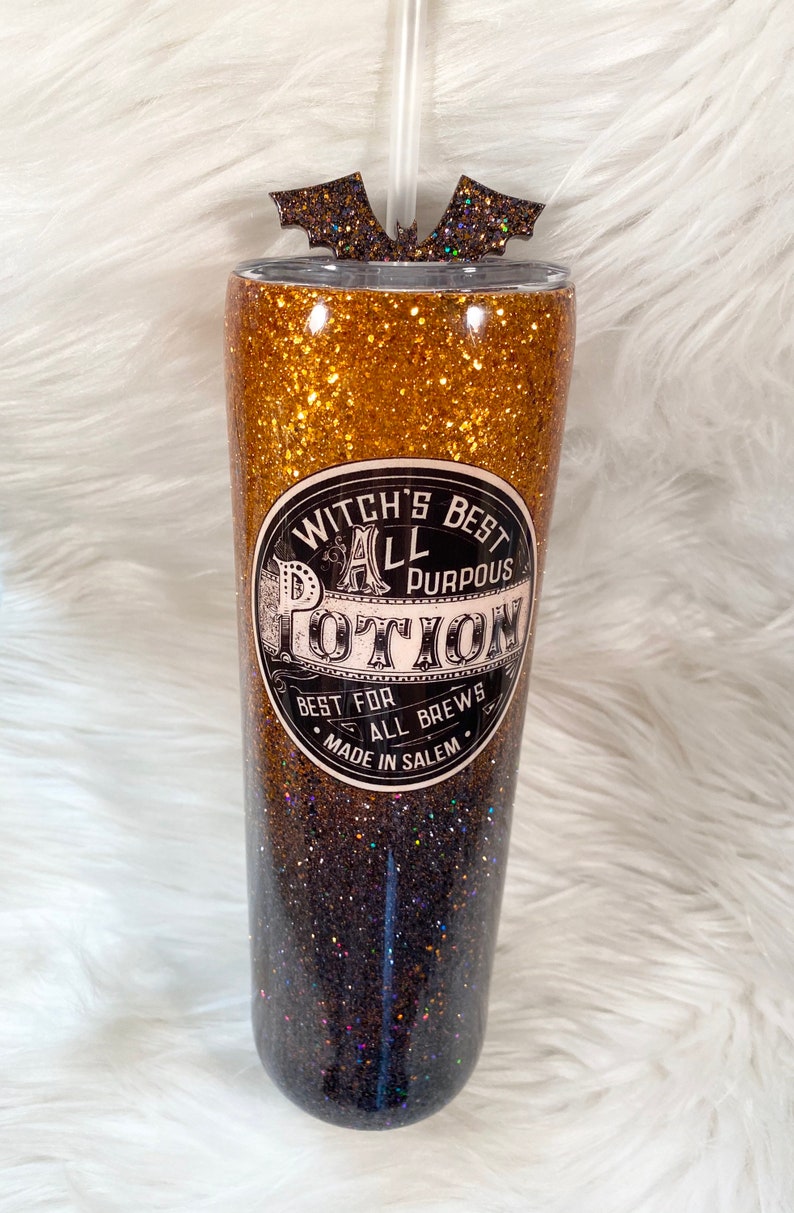 May include: A glitter-covered tumbler with a straw and bat-shaped topper. The tumbler features a gold and black ombre design and a label that reads "Witch's Best All Purpous Potion". The text "Best for All Brews, Made in Salem" is also on the label.