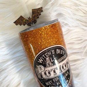 May include: A glittery gold and black tumbler with a bat-shaped straw topper. The tumbler features the text "Witch's Best All Purpose Potion" on a black and white label. A clear straw is inserted into the tumbler.