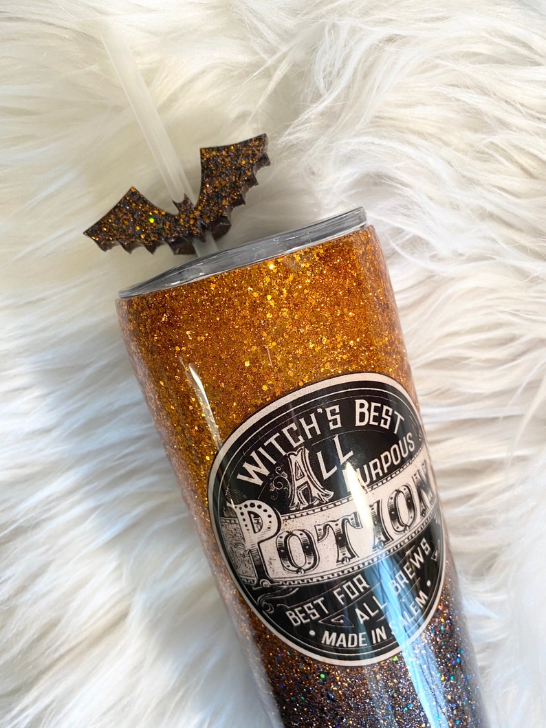 May include: A glittery gold and black tumbler with a bat-shaped straw topper. The tumbler features the text "Witch's Best All Purpose Potion" on a black and white label. A clear straw is inserted into the tumbler.