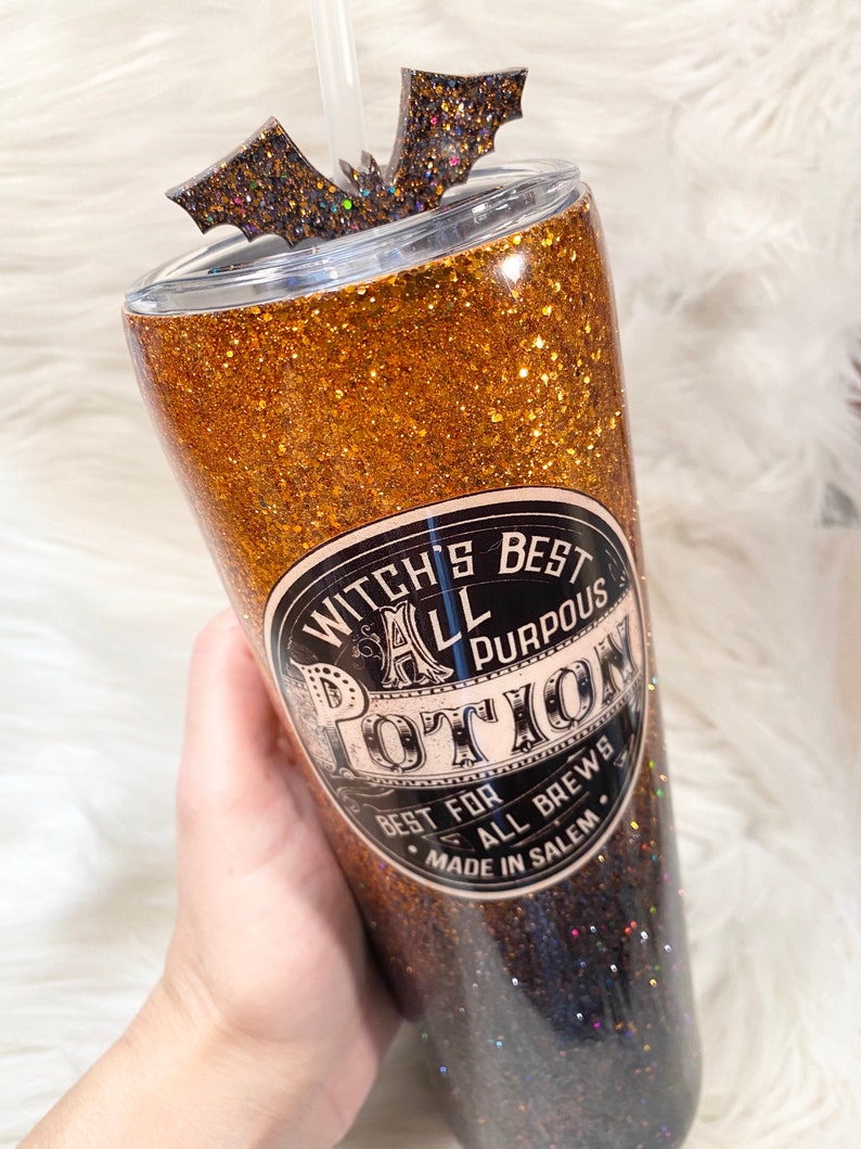 May include: A glittery tumbler with a bat-shaped topper and a straw. The tumbler is gold and black with the text "Witch's Best All Purpous Potion". The design is perfect for Halloween or a witchy theme.