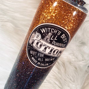 May include: A glittery tumbler with a gradient design, transitioning from gold to black. The tumbler features a label that reads "Witch's Best All Purpose Potion" and includes a straw and decorative accents on the lid. The tumbler is designed for cold beverages.