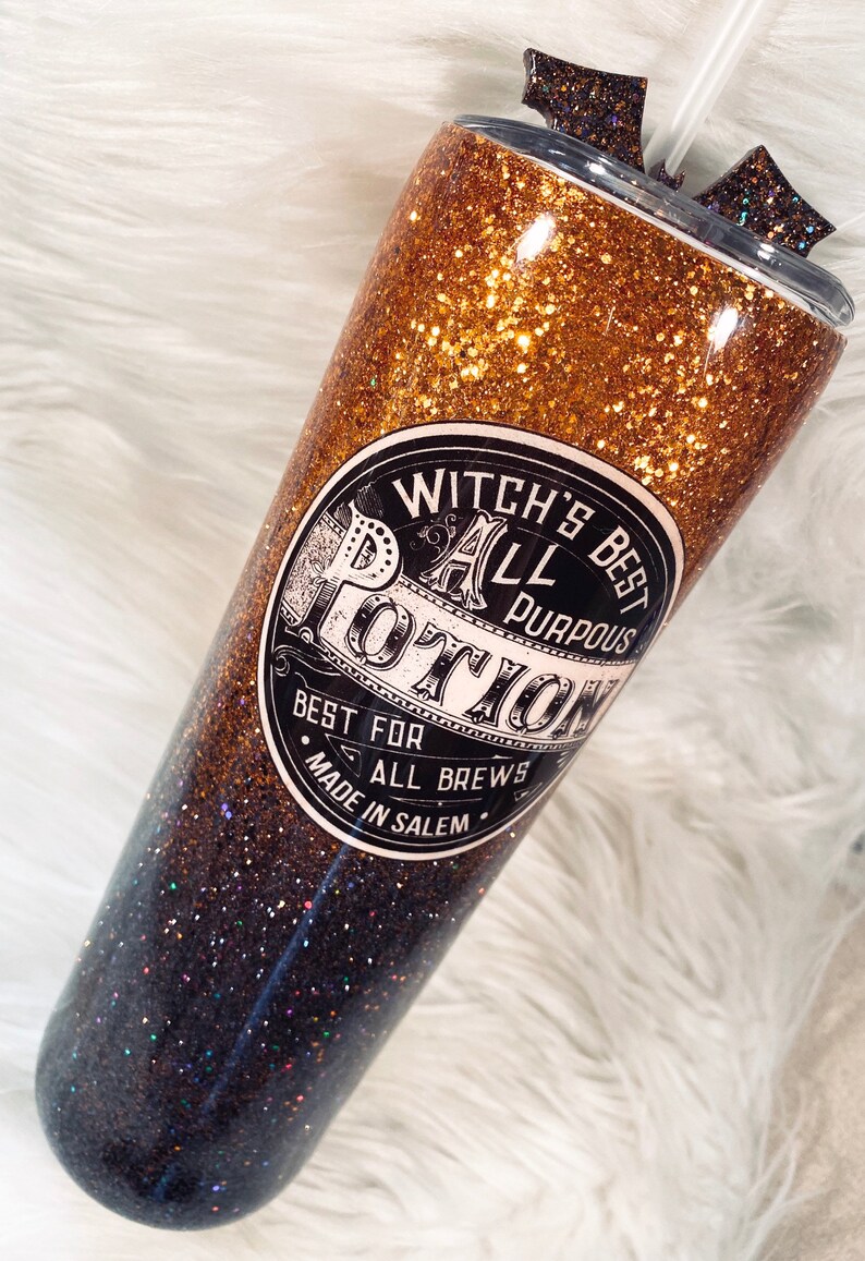 May include: A glittery tumbler with a gradient design, transitioning from gold to black. The tumbler features a label that reads "Witch's Best All Purpose Potion" and includes a straw and decorative accents on the lid. The tumbler is designed for cold beverages.