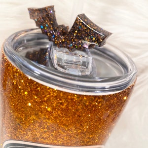 May include: A glittery gold tumbler with a black bat-shaped lid accent. The tumbler features the text "Witch's Best". A clear straw is inserted into the lid. The tumbler is designed for cold beverages.