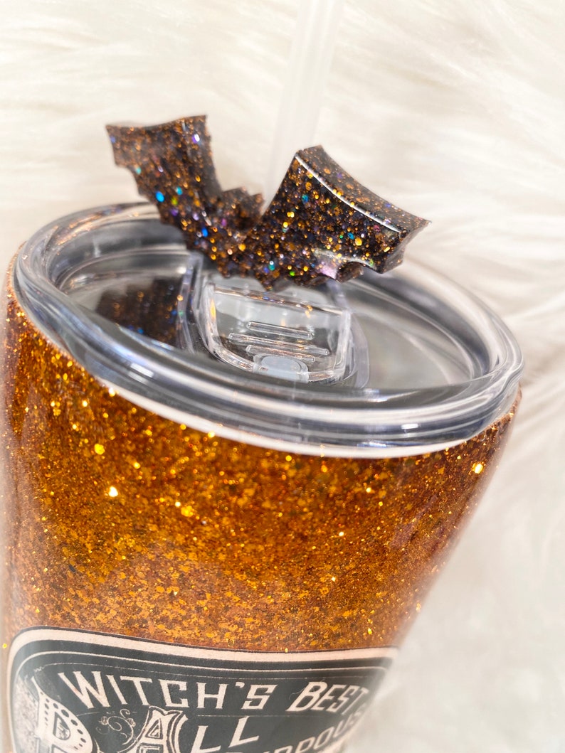 May include: A glittery gold tumbler with a black bat-shaped lid accent. The tumbler features the text "Witch's Best". A clear straw is inserted into the lid. The tumbler is designed for cold beverages.