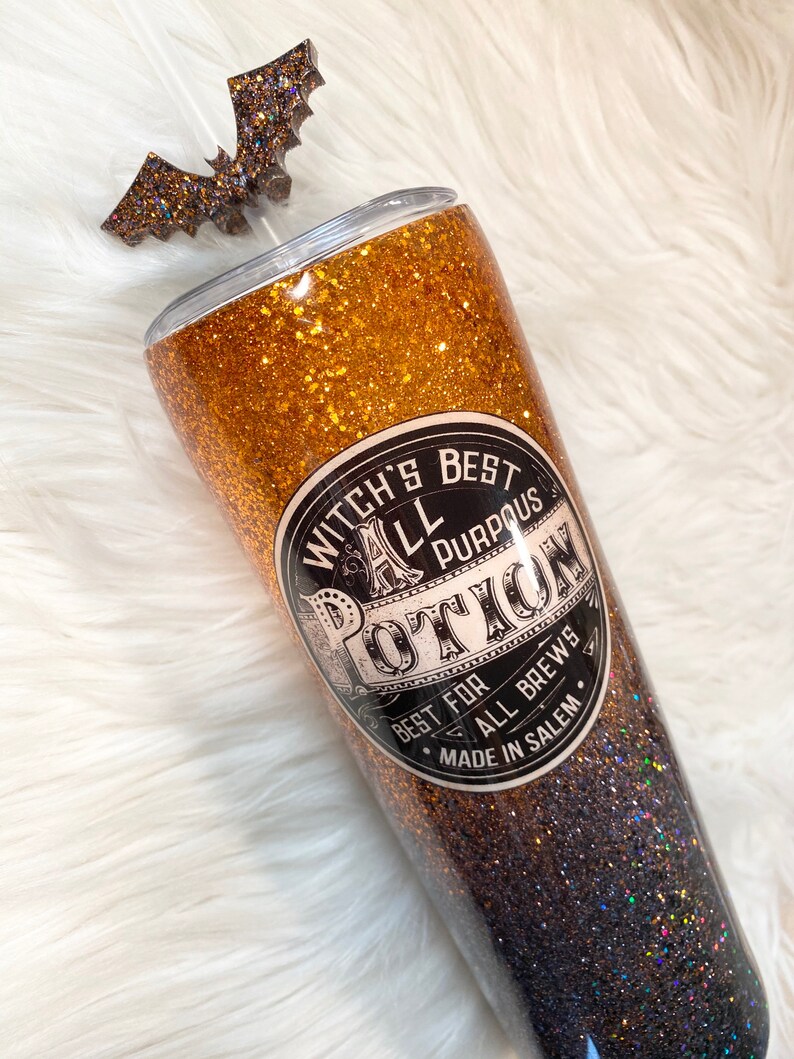 May include: A glittery tumbler with a bat-shaped topper. The tumbler is gold and black with the text "Witch's Best All Purpous Potion" on the front. The tumbler is made in Salem.