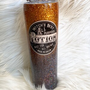 May include: A glittery tumbler with a gradient of gold to dark brown. The tumbler features a label that reads "Witch's Best All Purpous Potion" and a bat-shaped lid decoration. A clear straw is inserted.