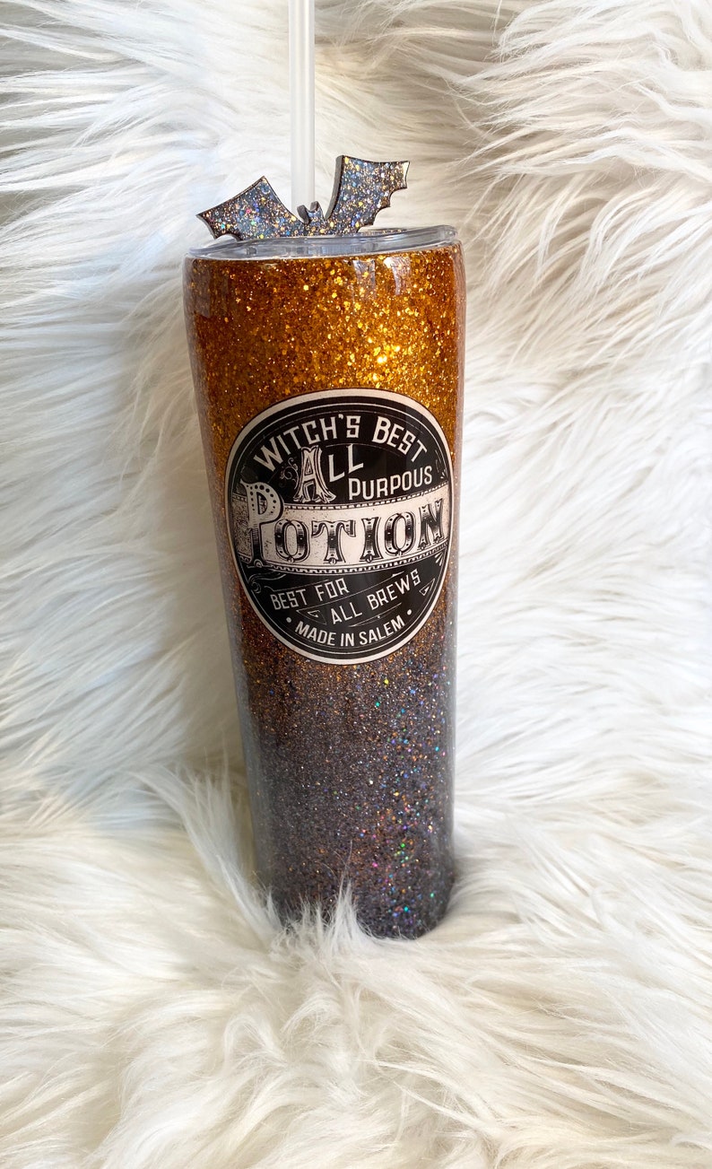May include: A glittery tumbler with a gradient of gold to dark brown. The tumbler features a label that reads "Witch's Best All Purpous Potion" and a bat-shaped lid decoration. A clear straw is inserted.