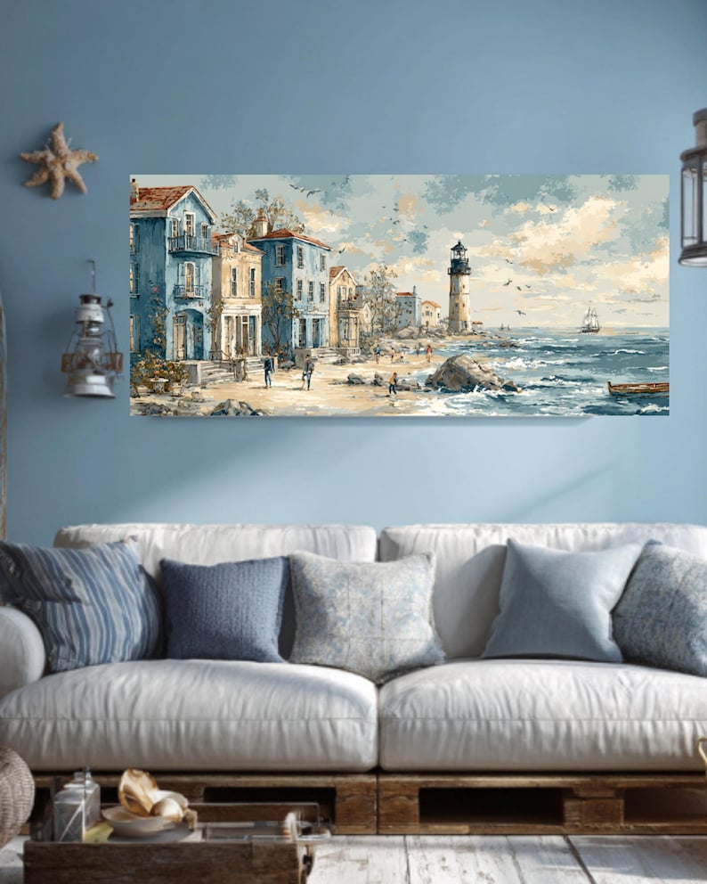 May include: A coastal landscape painting featuring buildings in shades of blue and yellow, a lighthouse, and a sailboat on the sea. The artwork depicts a beach scene with figures, birds, and a small boat. The painting is displayed above a white sofa with blue pillows.
