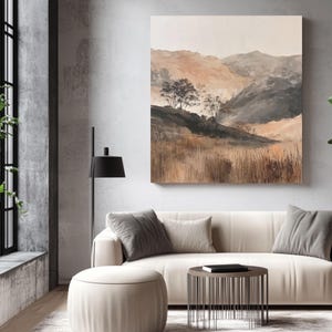 Puede incluir: Una impresión de arte mural cuadrada que presenta un paisaje de colinas onduladas, hierbas secas y árboles dispersos en tonos apagados de beige, marrón y gris carbón. La obra de arte se exhibe en una pared gris claro sobre un sofá color crema.