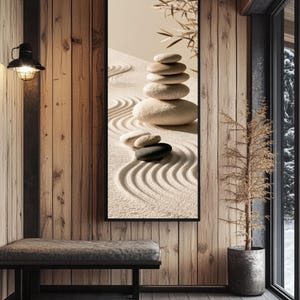 May include: A vertical rectangular wall art piece featuring a stack of five smooth, light-colored stones resting on a bed of raked sand with wavy patterns. A small dark stone sits nearby, and a bamboo branch extends from the top right corner.