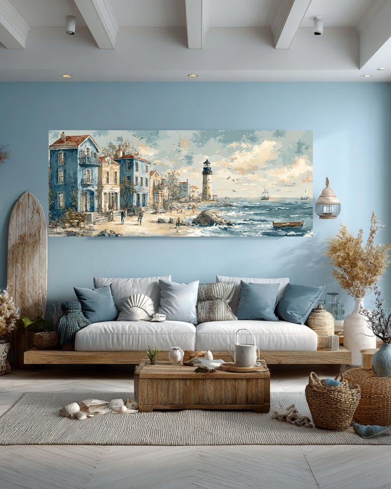 May include: Coastal-themed artwork depicting a seaside town with blue buildings, a lighthouse, and a beach. The painting is displayed above a white sofa with blue pillows, complemented by a wooden coffee table and decorative elements, creating a serene living room setting.