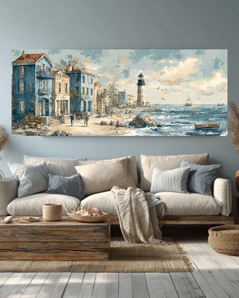 May include: A coastal landscape painting depicts buildings in shades of blue and yellow along a beach, with a lighthouse in the distance. The artwork features a serene ocean with small boats and a cloudy sky. The painting is displayed above a beige sofa with decorative pillows.