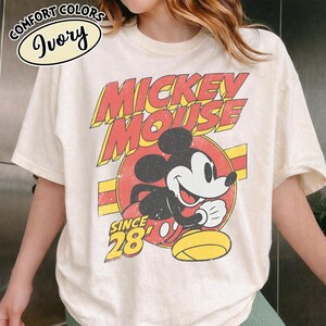 以下が含まれることがあります： アイボリー色のTシャツ。ミッキーマウスのヴィンテージ風グラフィックが特徴です。「Mickey Mouse」の文字が赤と黄色で描かれ、その下に「Since 28」とあります。Comfort Colorsブランドのシャツです。