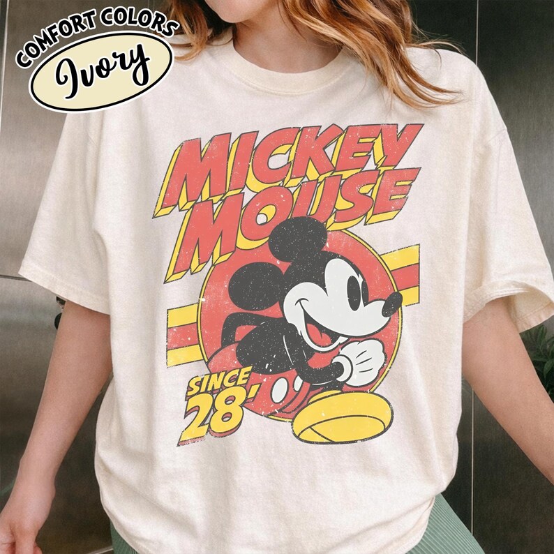 以下が含まれることがあります： アイボリー色のTシャツ。ミッキーマウスのヴィンテージ風グラフィックが特徴です。「Mickey Mouse」の文字が赤と黄色で描かれ、その下に「Since 28」とあります。Comfort Colorsブランドのシャツです。