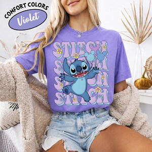 Puede incluir: Camiseta Comfort Colors violeta con un dise&ntilde;o de Stitch de Lilo & Stitch. El dise&ntilde;o incluye la palabra "Stitch" en una fuente retro con detalles florales. La camiseta es de color morado claro.