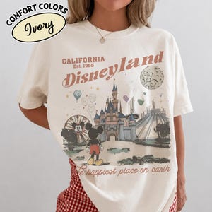 May include: Ivory-colored t-shirt with a Disneyland graphic. The design features a castle, Mickey Mouse, a Ferris wheel, and the text "California Disneyland Est. 1955." The phrase "The happiest place on earth" is also included.