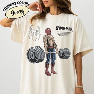 May include: Ivory-colored t-shirt with a Spider-Man graphic. The design features a weightlifting Spider-Man with tattoos, a spider symbol, and the text "SPIDER-MAN". The shirt is labeled "Comfort Colors".
