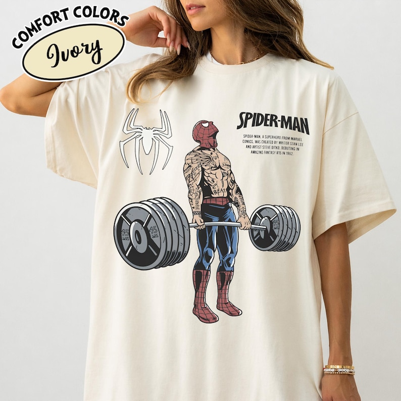 May include: Ivory-colored t-shirt with a Spider-Man graphic. The design features a weightlifting Spider-Man with tattoos, a spider symbol, and the text "SPIDER-MAN". The shirt is labeled "Comfort Colors".