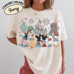 以下が含まれることがあります： アイボリー色のTシャツ。カラフルなグラフィックには、漫画のキャラクターと象徴的なランドマークが描かれています。デザインには花火、お城、自由の女神が含まれています。「Comfort Colors」と「Ivory」の文字がプリントされています。