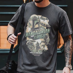 May include: Dark gray t-shirt featuring a graphic from the Star Wars series, "The Mandalorian and Grogu." The design includes the Mandalorian helmet, Grogu, and the phrase "This is the Way." The shirt has a vintage, worn look.