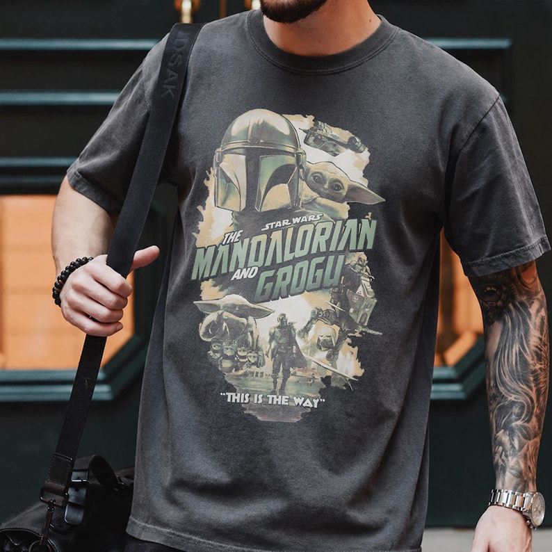 May include: Dark gray t-shirt featuring a graphic from the Star Wars series, "The Mandalorian and Grogu." The design includes the Mandalorian helmet, Grogu, and the phrase "This is the Way." The shirt has a vintage, worn look.